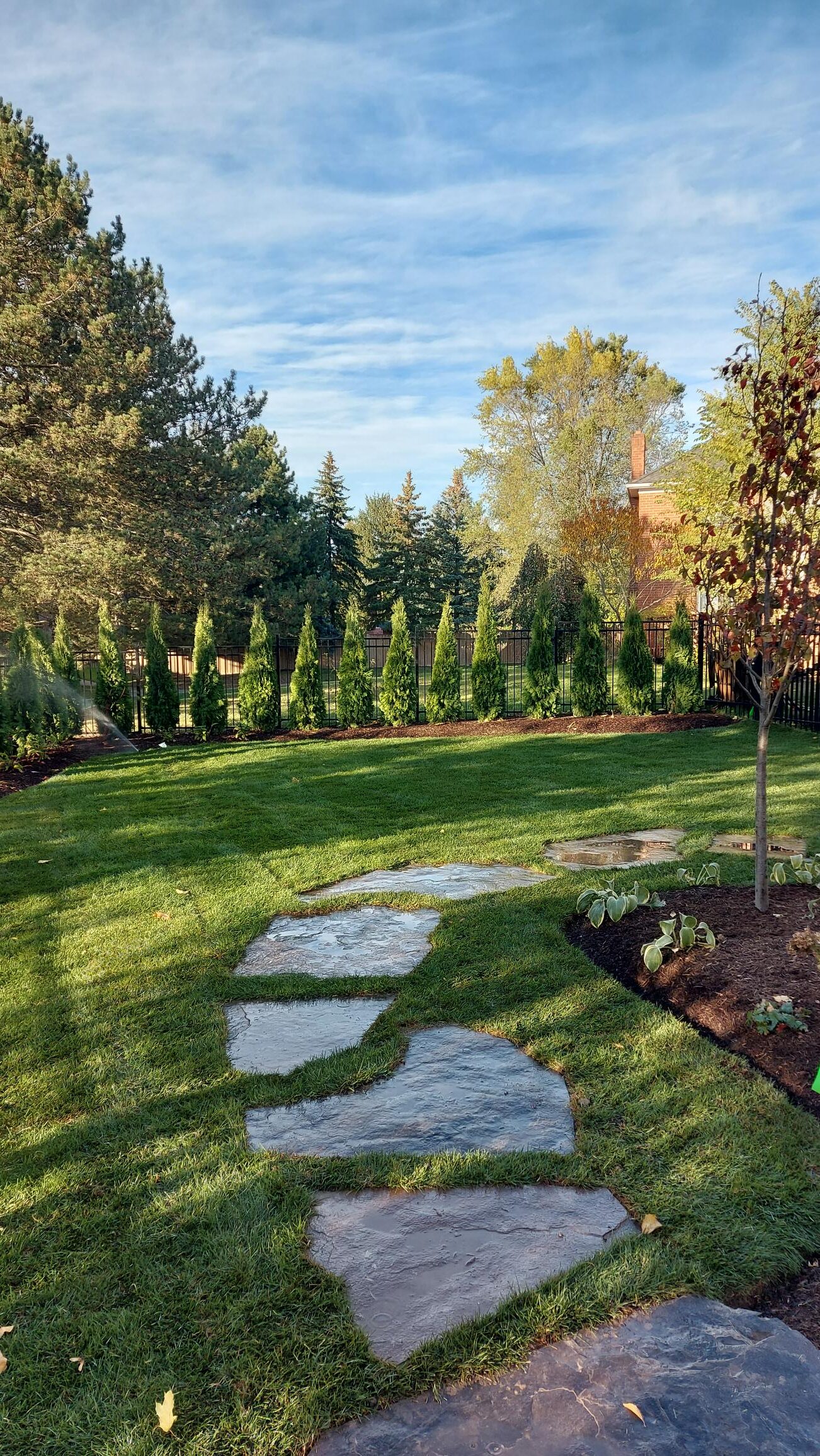 A peaceful garden with a stone path, lush grass, trees, and a red-brick house partially visible in the background under a blue sky.
