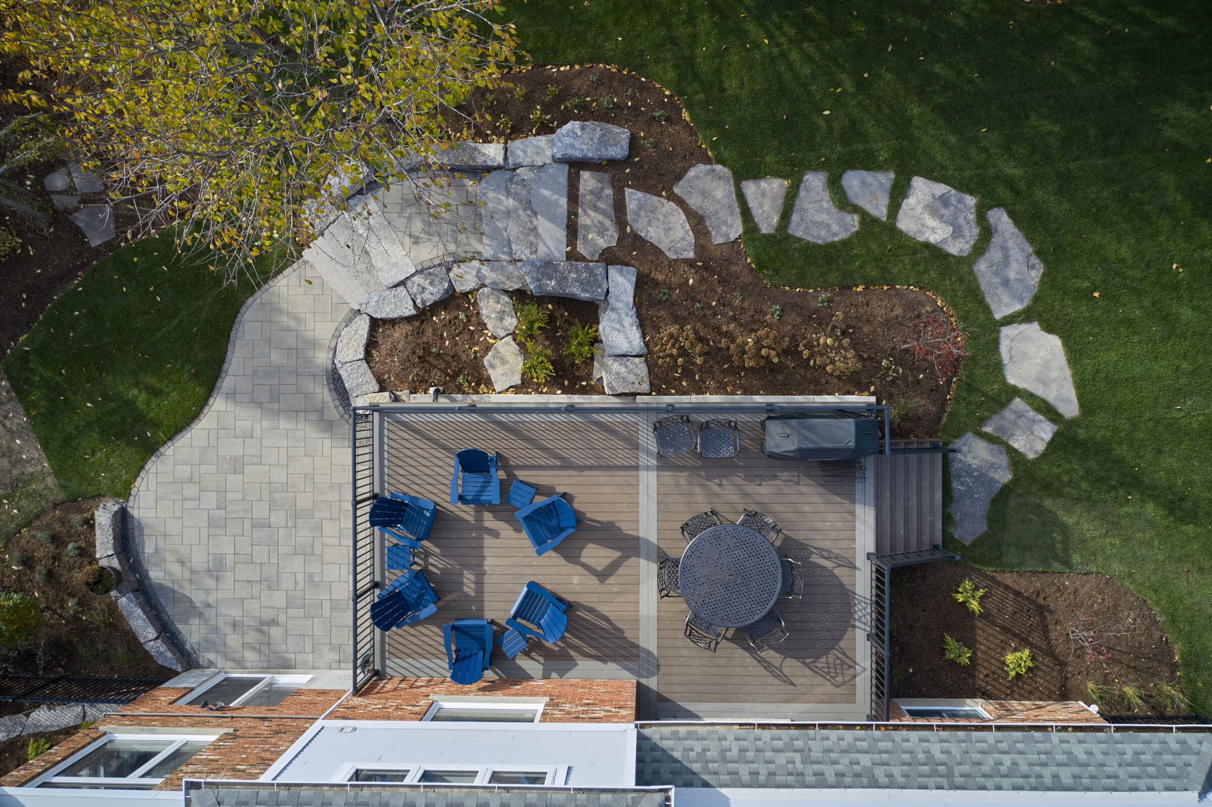 Aerial view of a backyard patio with blue chairs, round table, and stone pathway surrounded by grass and garden landscaping.