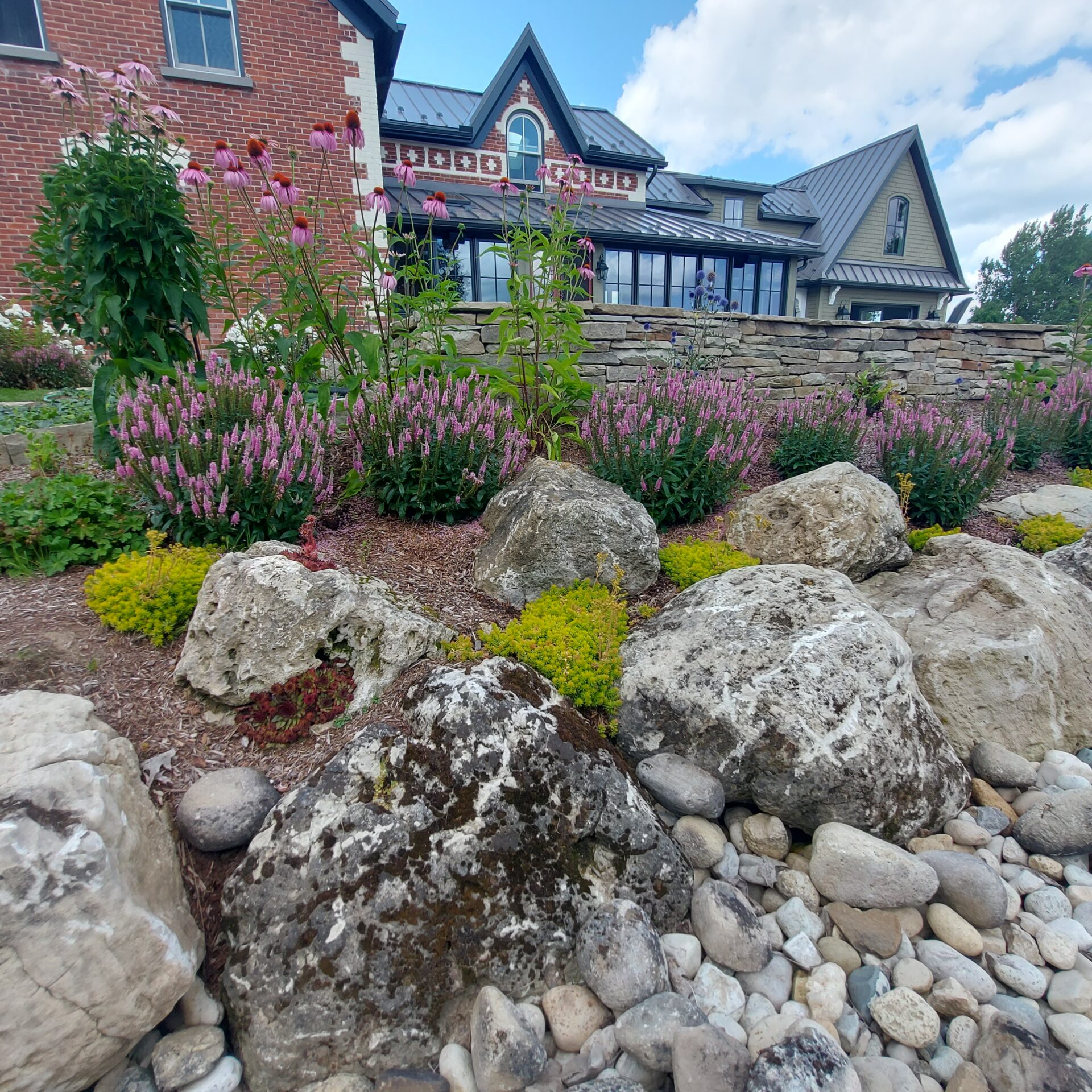 A charming garden with rocks, colorful flowers, and a stone house featuring a gabled roof, set under a partly cloudy sky.