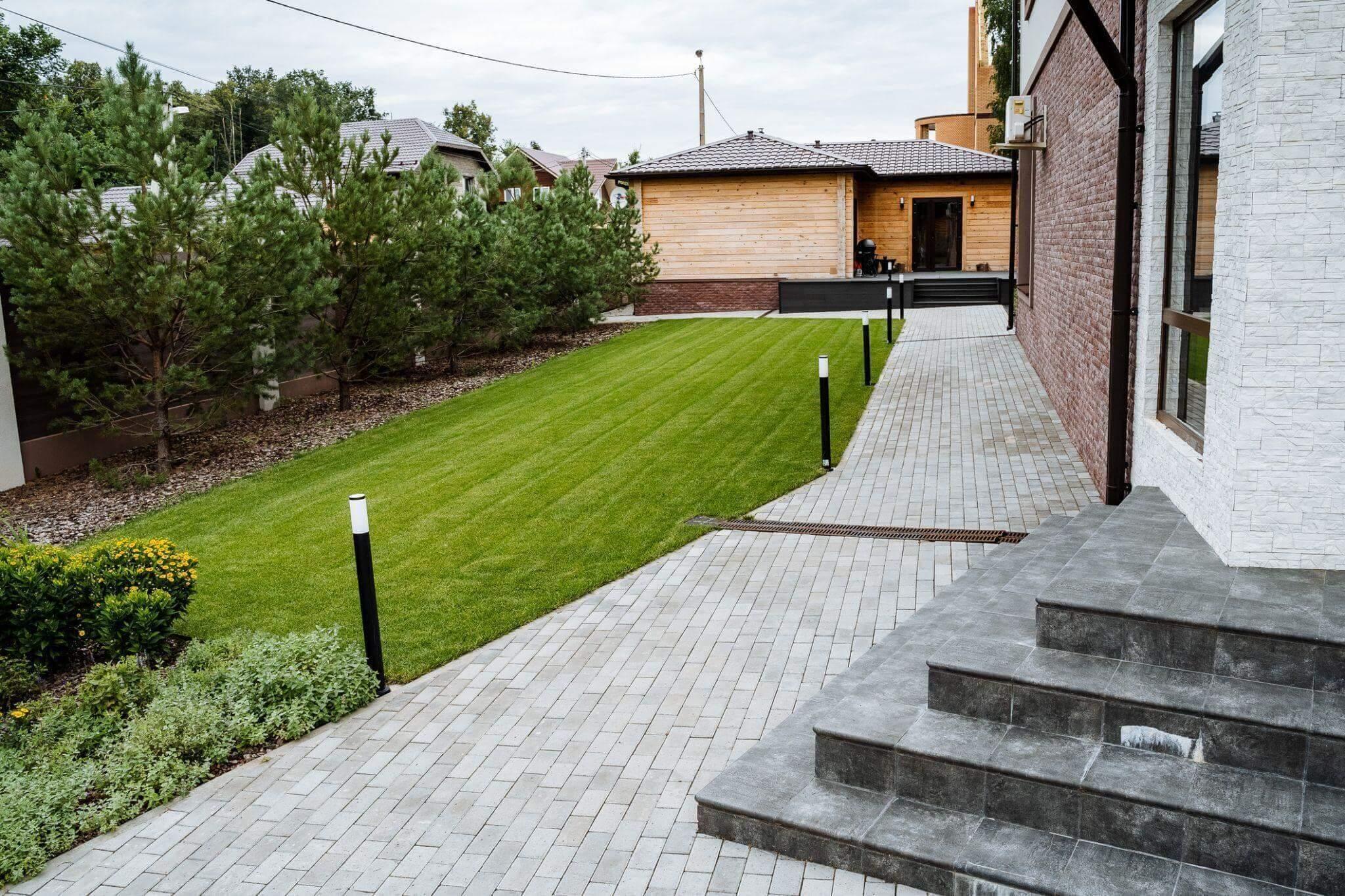 A manicured lawn leads to a small wooden building, surrounded by trees and paved pathways with steps near a modern house.