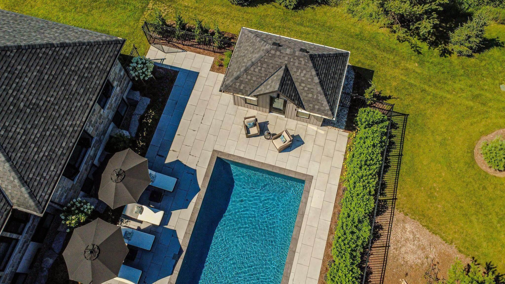 Aerial view of a swimming pool, surrounded by patio seating and green lawn, adjacent to a large building with umbrellas and lounge chairs.