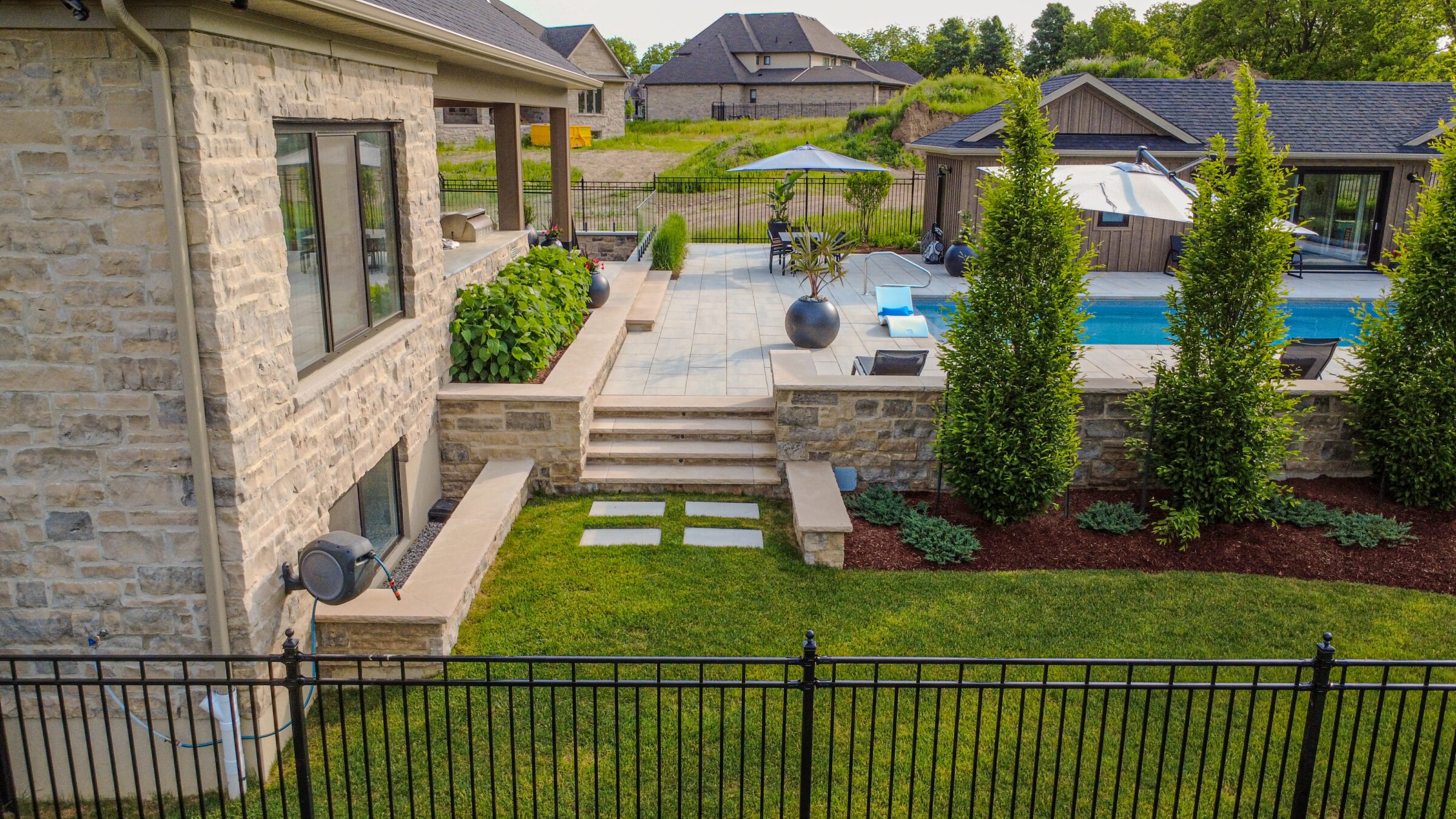 Modern backyard with stone patio, pool, and lounge chairs. Lush greenery surrounds stone steps. Fenced area with well-maintained lawn and ornamental trees.