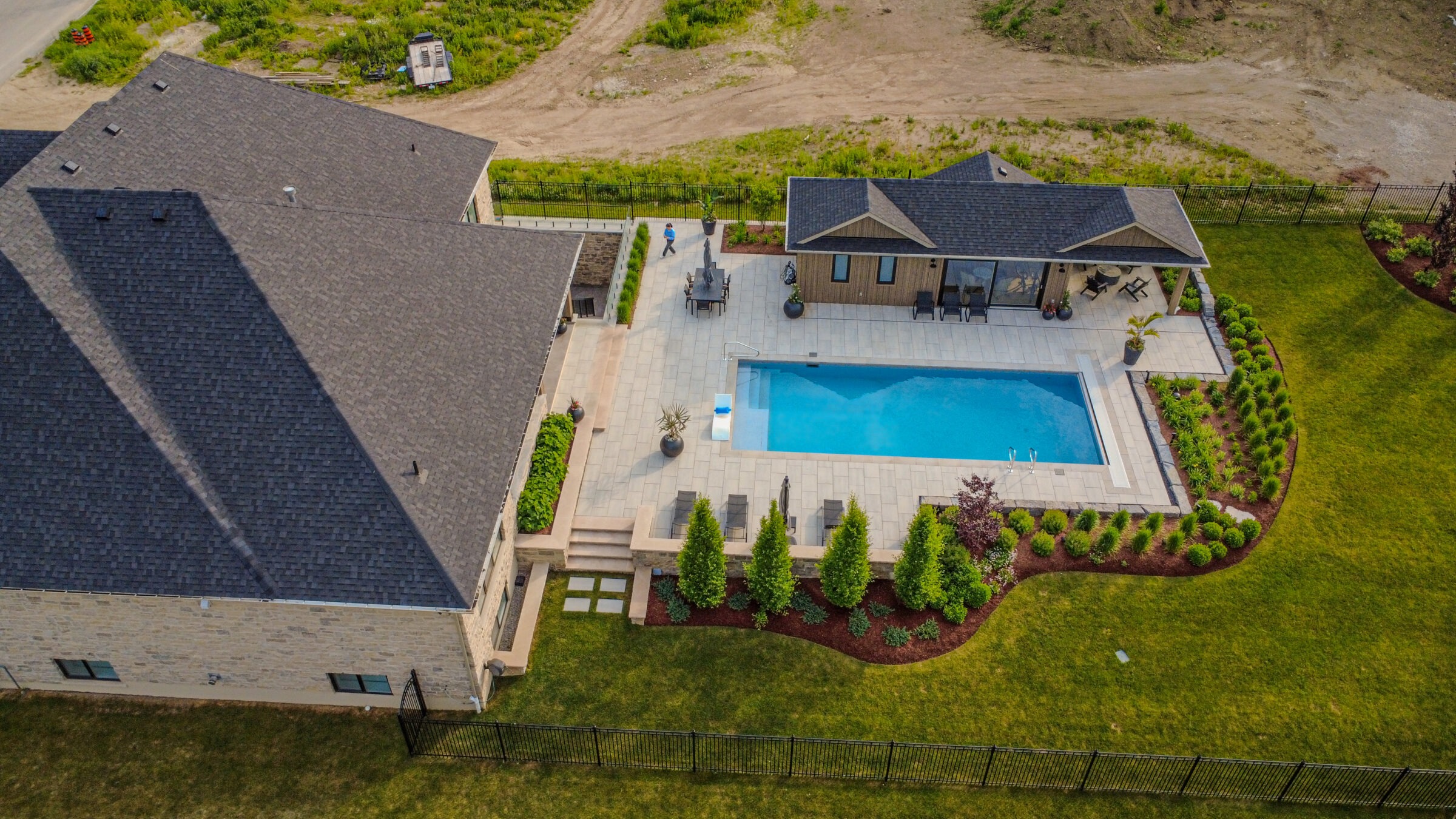 Aerial view of a backyard with a modern house, swimming pool, landscaped garden, patio furniture, and a person walking nearby.