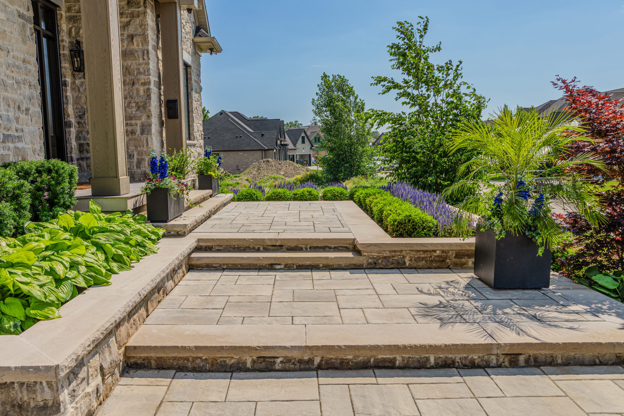 Elegant stone patio with lush greenery and vibrant flowers, leading to a suburban home. Clear blue sky and neighboring houses visible.