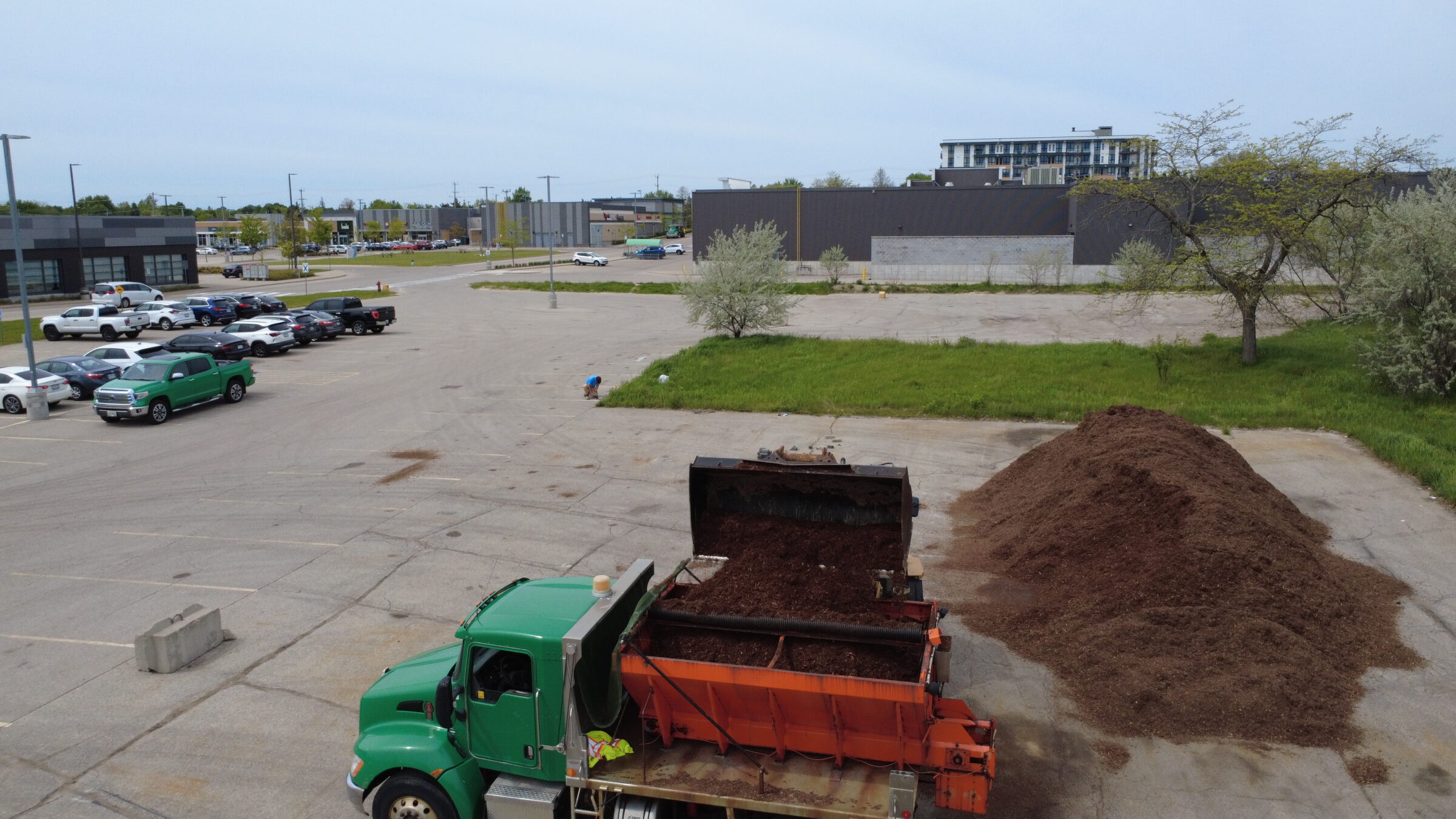 A green truck unloads soil in a parking lot. Nearby, a person kneels on grass, with parked cars and buildings in the background.