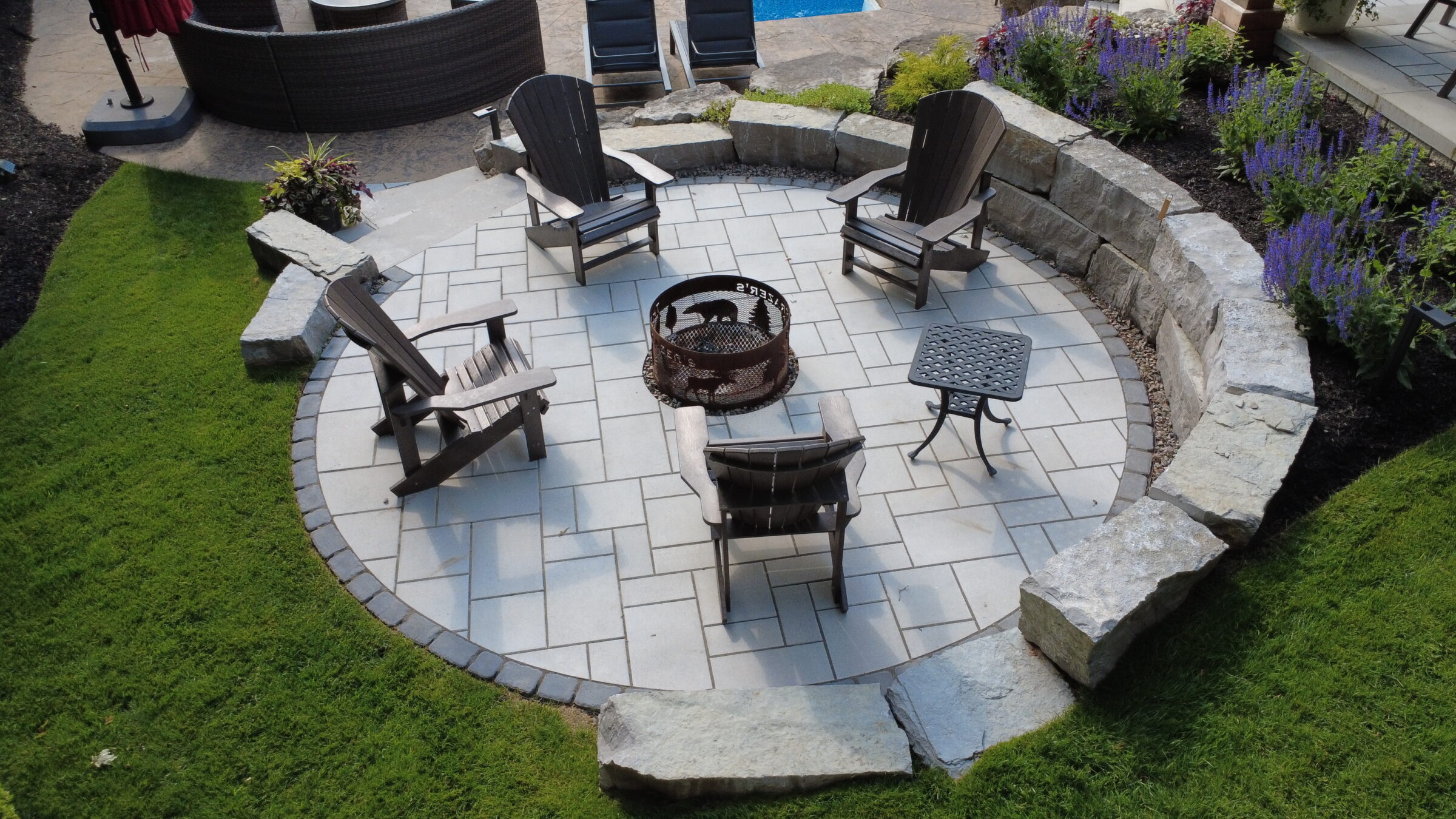 A stone patio features Adirondack chairs around a fire pit, surrounded by lush grass, colorful flowers, and patio furniture.