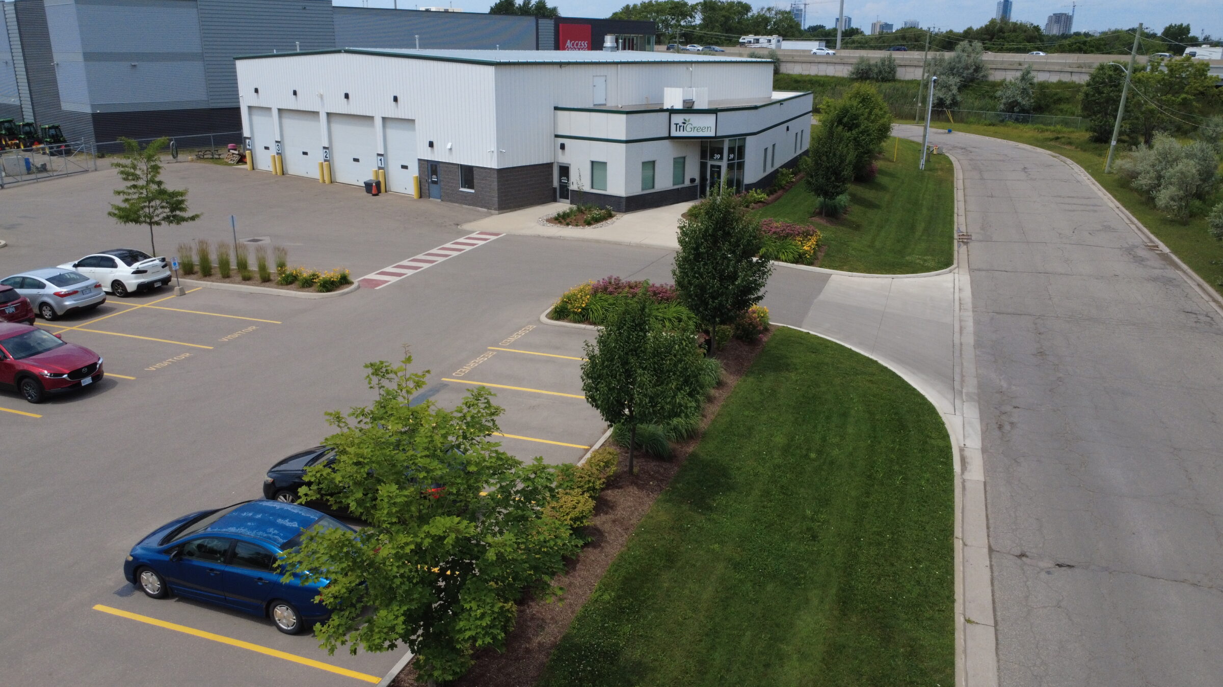 Industrial building with parking lot, surrounded by greenery. Several cars parked. Curved road and highway in background under a clear sky.