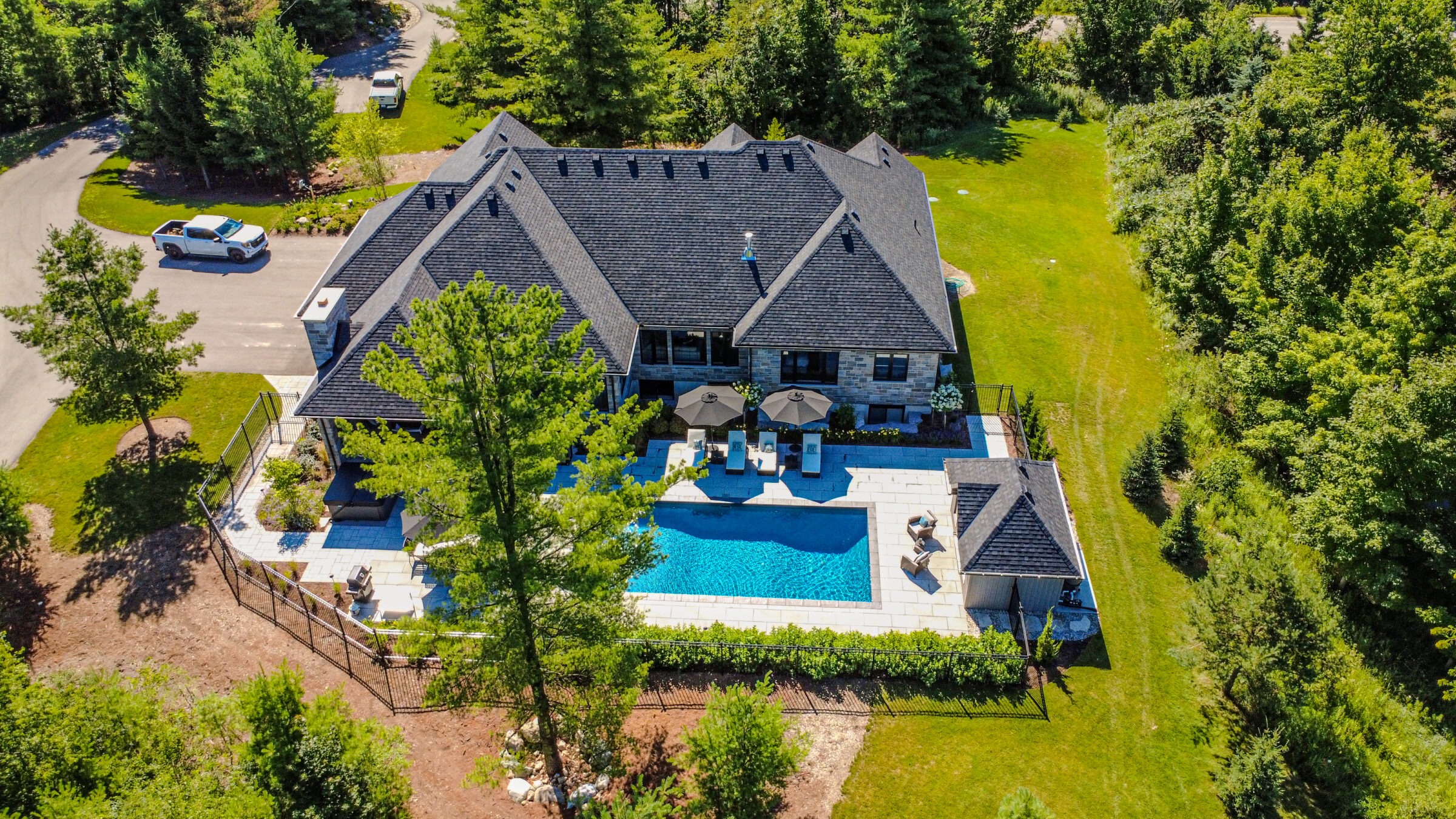 Luxurious house surrounded by trees, features a large swimming pool, patio with umbrellas, and parked cars on a nearby road.
