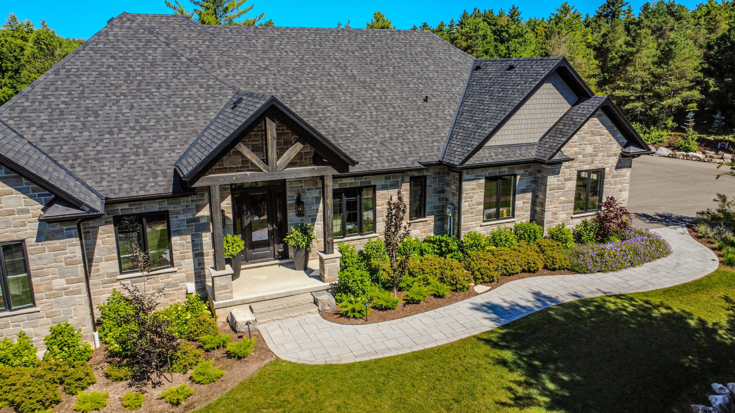 A modern stone house with a dark shingled roof, surrounded by well-maintained gardens and a paved walkway, set against lush greenery.