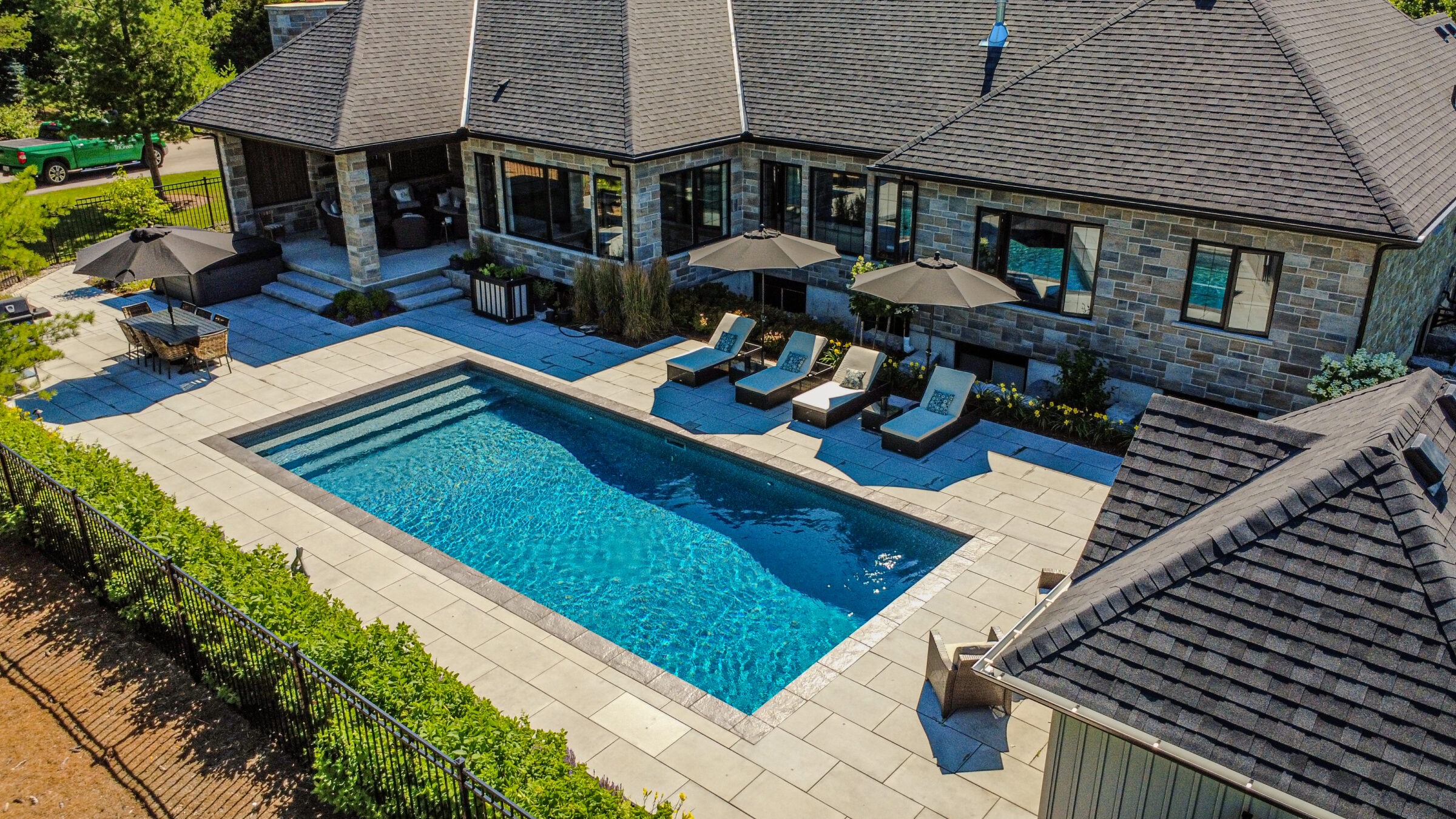 A stone house with a patio features a swimming pool, lounge chairs, umbrellas, and outdoor dining furniture, surrounded by greenery and pavement.