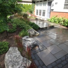 A well-maintained garden features a stone pathway, shrubs, and rocks, leading to a house with brick and siding exterior and large windows.