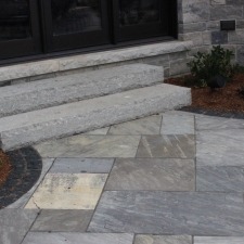 Stone steps lead to a modern building entrance. The patio features gray tiles and minimal landscaping with small shrubs by the doorway.