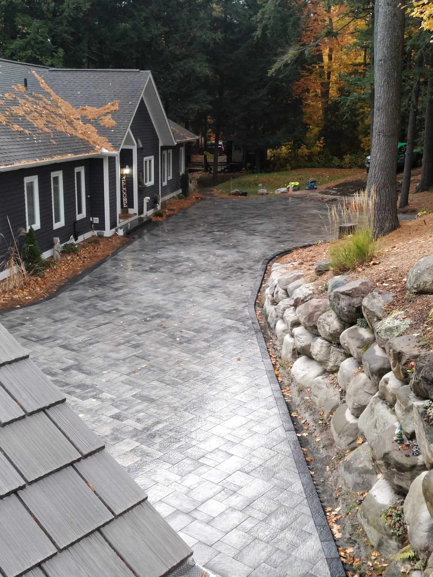 A modern house with a paved driveway, surrounded by autumn leaves and tall trees, offers a peaceful, secluded forest setting.