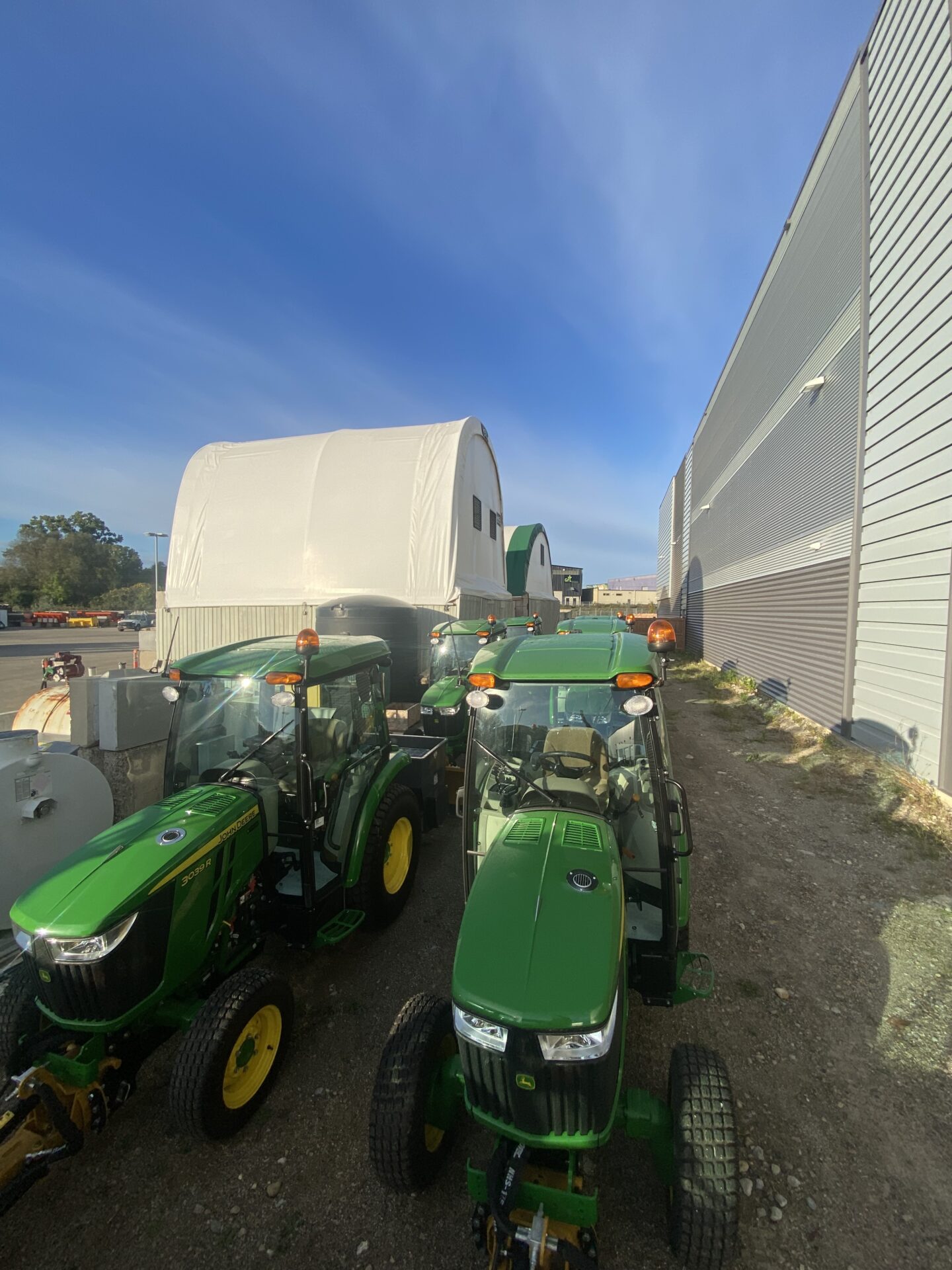 Tractors are parked near industrial buildings under a clear blue sky, with a large white tent structure visible in the background.