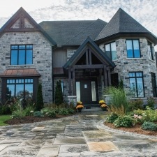 A large stone house with a steep roof, multiple windows, and lush landscaping. A stone pathway leads to the entrance.