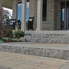 A stone-paved entrance features stairs leading to a porch with columns, chairs, and plants, outside a modern brick house.