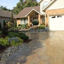 Suburban house with stone pathway, landscaped garden, and modern architecture. Features a well-maintained driveway and lush greenery, under a clear sky.