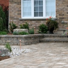 Brick house with white windows, stone-paved patio, and neatly arranged garden featuring potted plants and decorative stones in the foreground.
