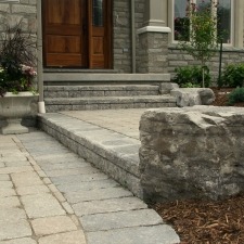 Stone steps lead to a wooden door of a residential building, surrounded by brick walls and small plants, with a paved pathway.