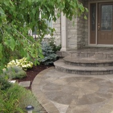 Stone pathway leads to a home's front entrance with circular steps, surrounded by lush greenery and a small garden under tree shade.
