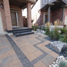 Brick houses with stone steps and landscaped front yard. Decorative pavers and plants are arranged alongside large rocks, creating a neat and welcoming entrance.