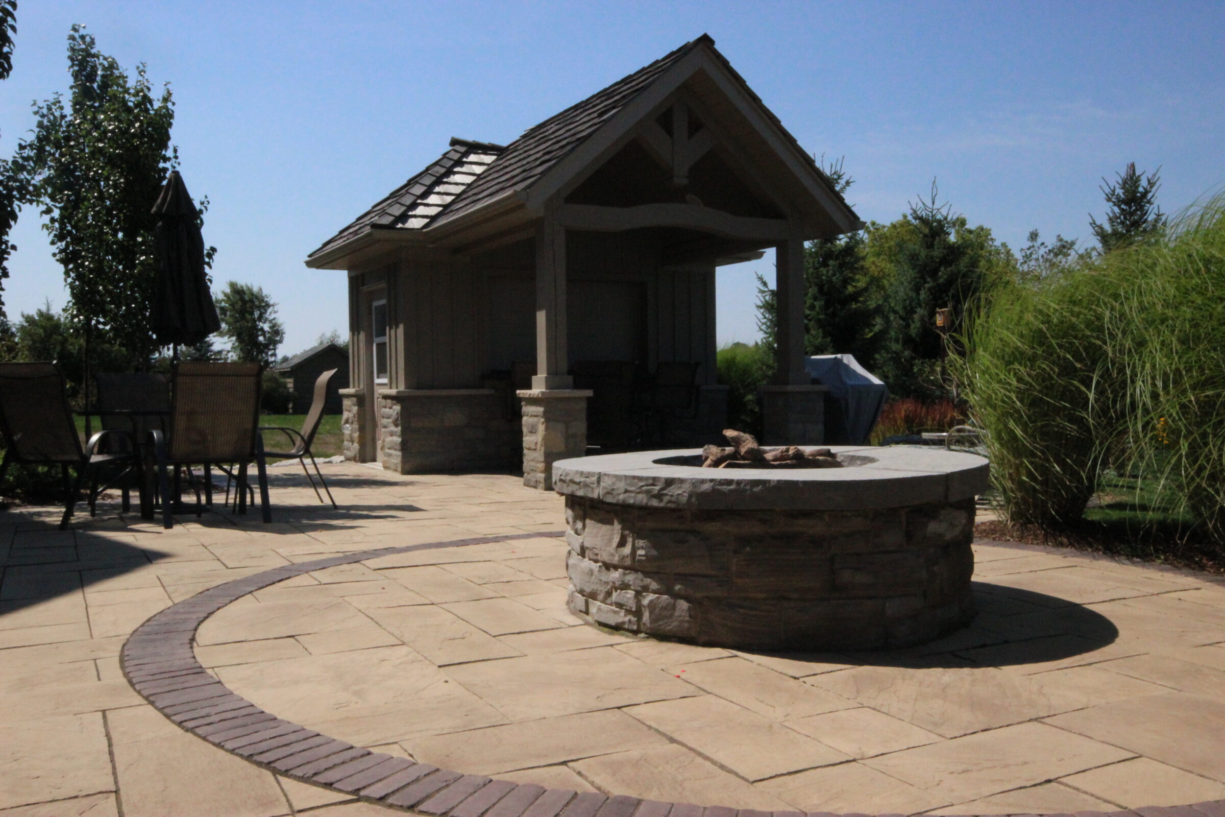 Outdoor patio features a fire pit, seating area, and small pavilion. Surrounded by greenery and trees under a clear blue sky.