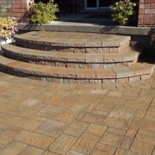 A set of semi-circular, stone steps leads to a brick building entrance, flanked by potted plants on a sunlit patio.
