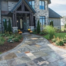 A stone pathway leads to a modern house with a gabled entrance, surrounded by landscaped gardens and decorative plants under a cloudy sky.