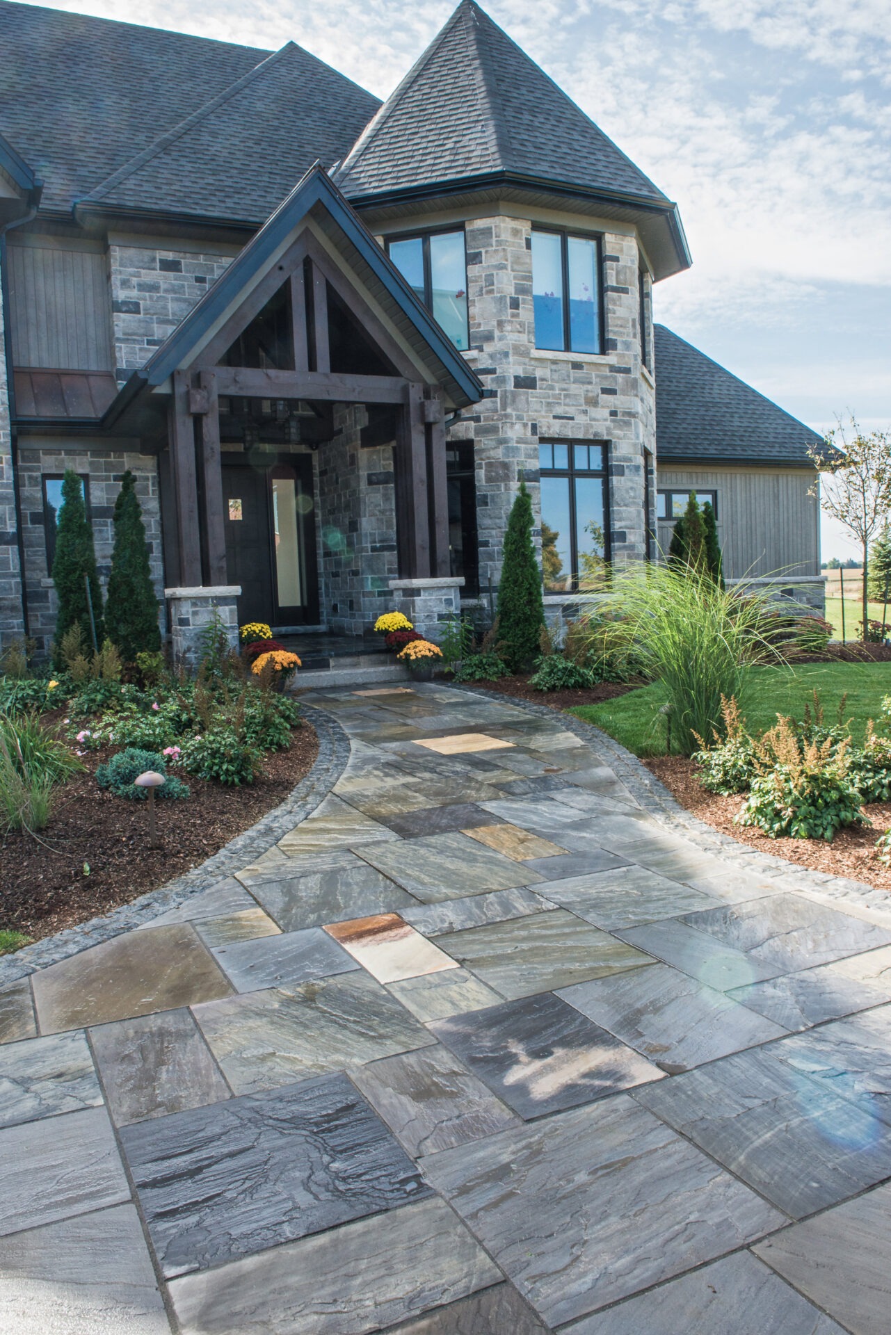 A stone house with a peaked roof, surrounded by a landscaped garden and a paved walkway under a clear blue sky.