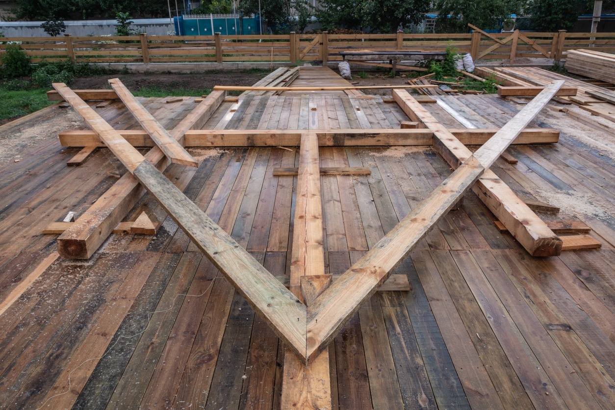Wooden beams arranged on a newly built deck, fenced area in the background, with green foliage surrounding the site.
