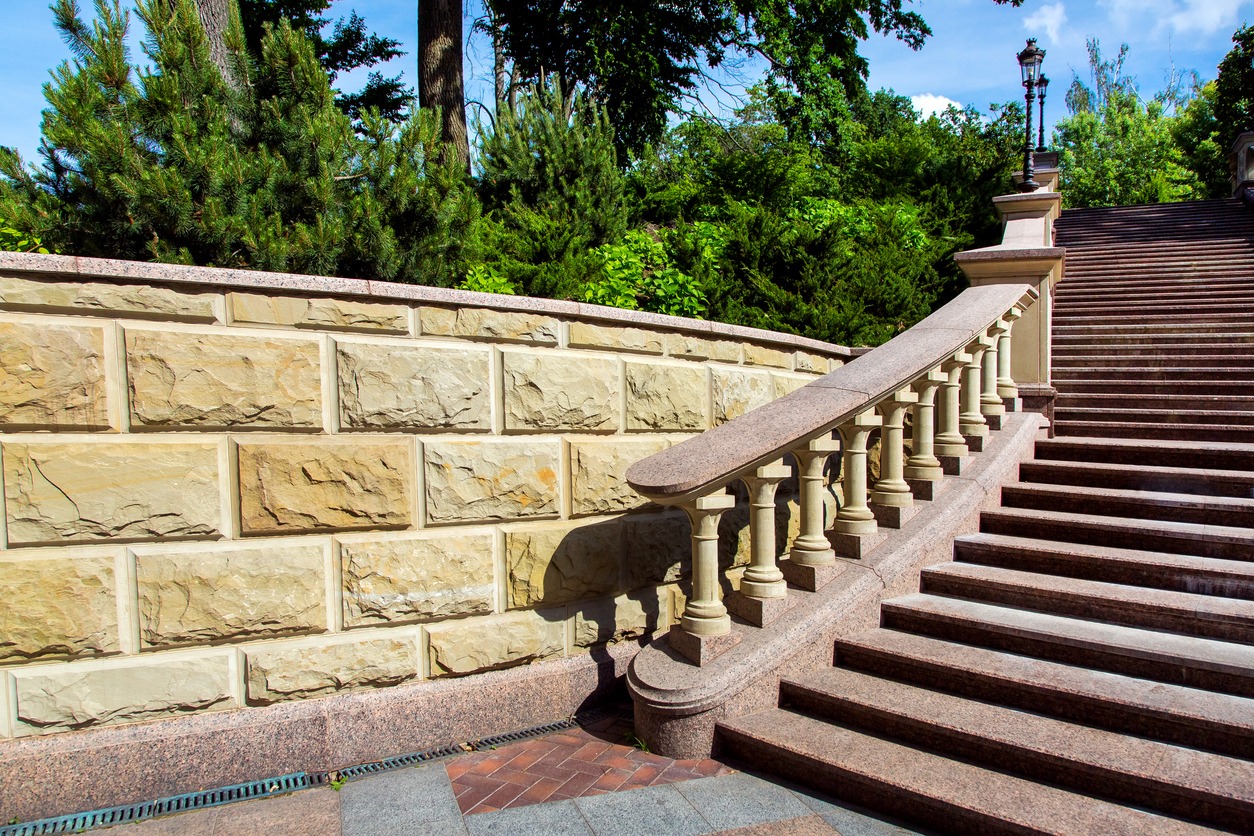 Stone staircase with balustrade leads through lush greenery under a blue sky. Sunlight casts shadows on the decorative stone wall.