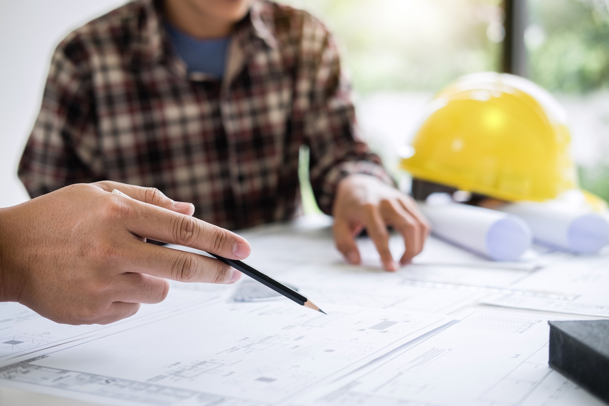 Two people reviewing architectural blueprints with pencils. A yellow hard hat and rolled documents are on the table, suggesting a construction project.