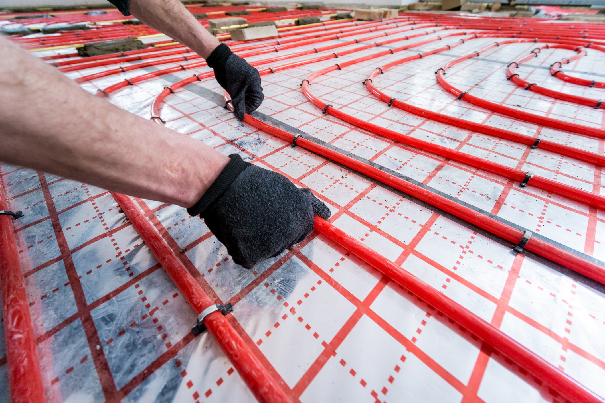 A person wearing gloves installs red pipes over a grid-patterned surface, likely for an underfloor heating system.