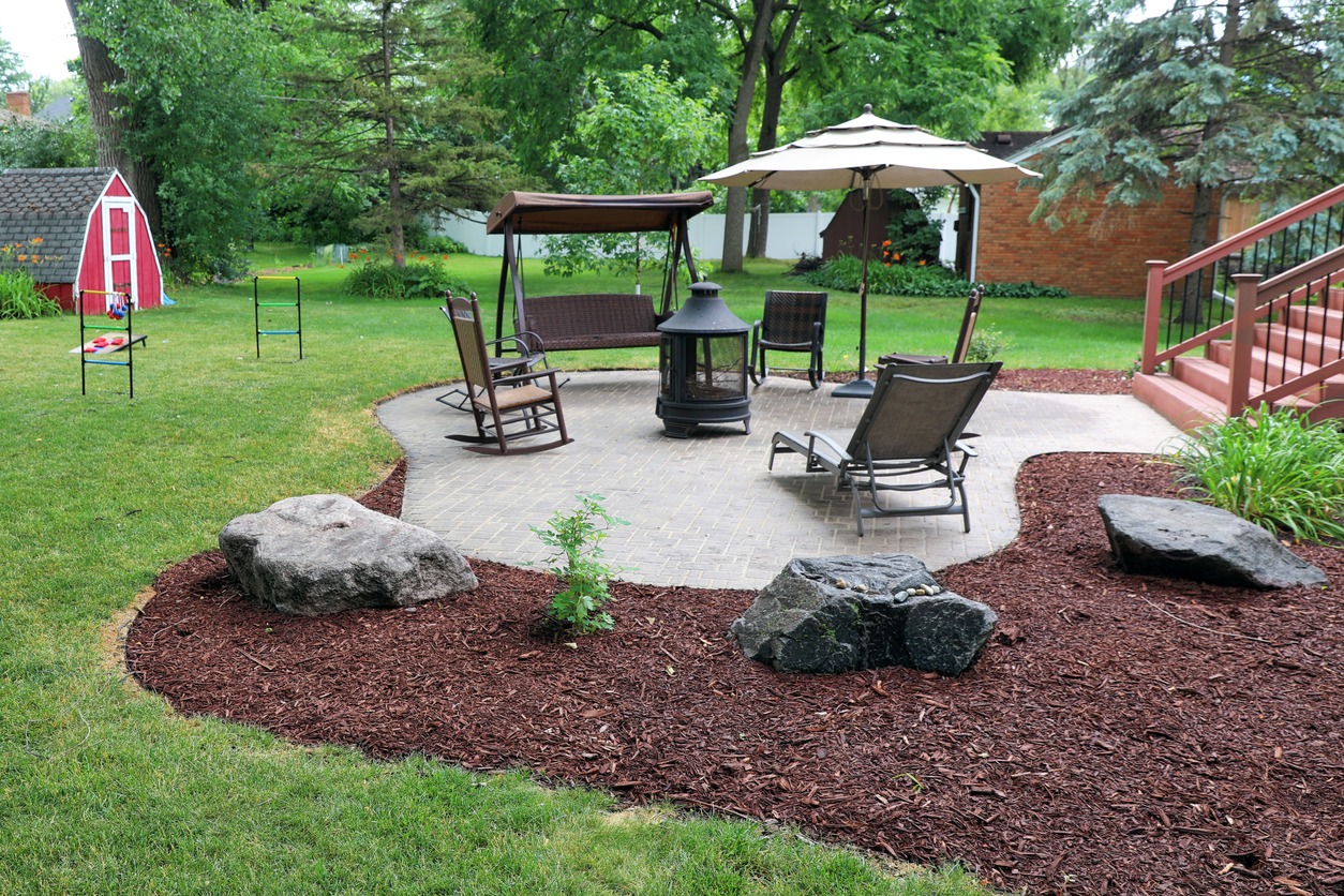 A serene backyard features patio furniture, a fire pit, a small red shed, lush greenery, and garden mulch around landscaped rocks.