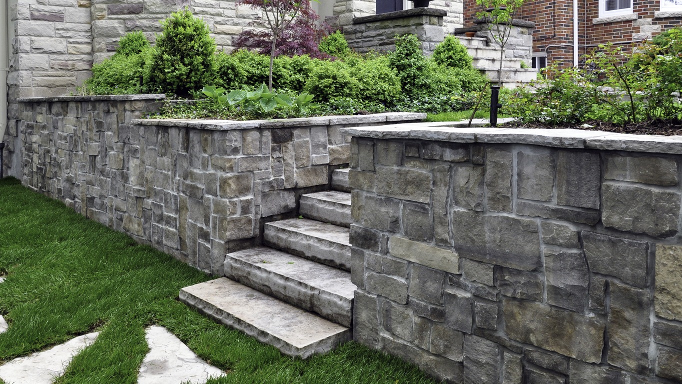 Stone steps lead through a manicured garden featuring lush greenery and a tree, surrounded by stone walls and brick building exteriors.