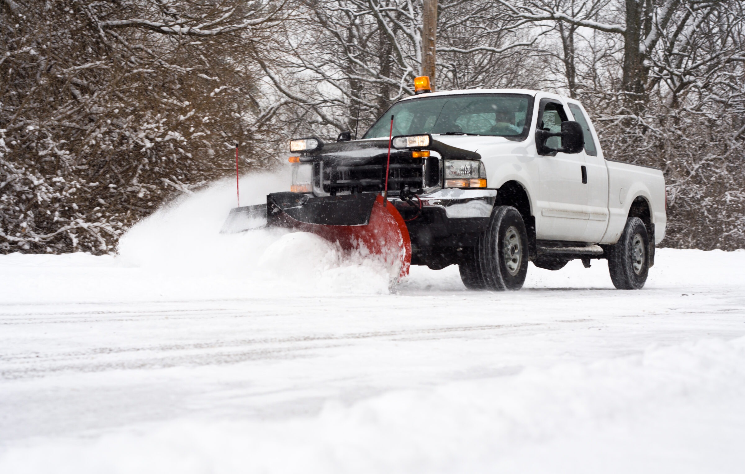 Truck with a snowplow clearing snowy road, surrounded by snow-covered trees. No recognizable landmarks or buildings are visible in the scene.