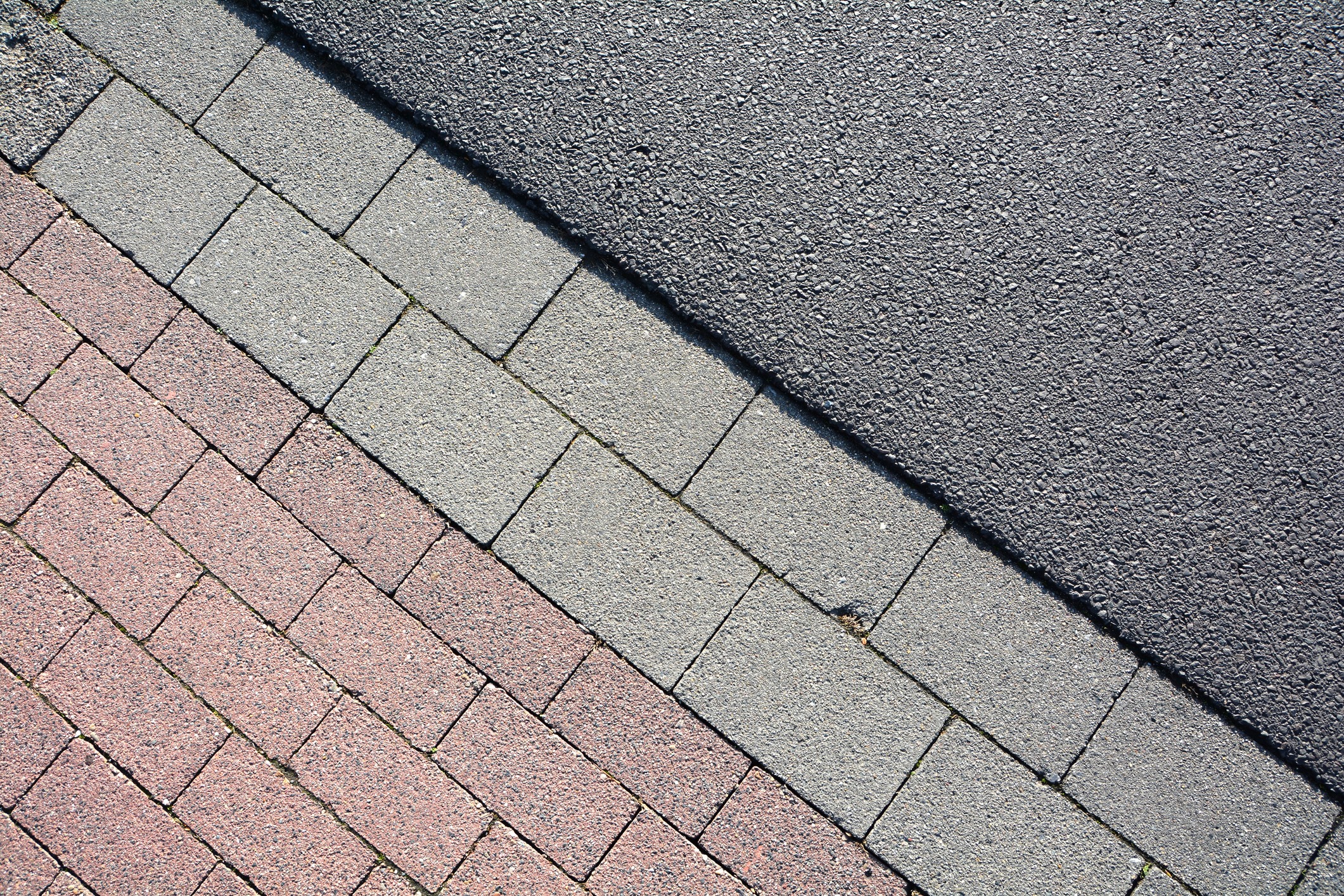 Diagonal view of a sidewalk showing red brick, gray stone, and asphalt sections, illustrating contrasting textures and colors in urban design.