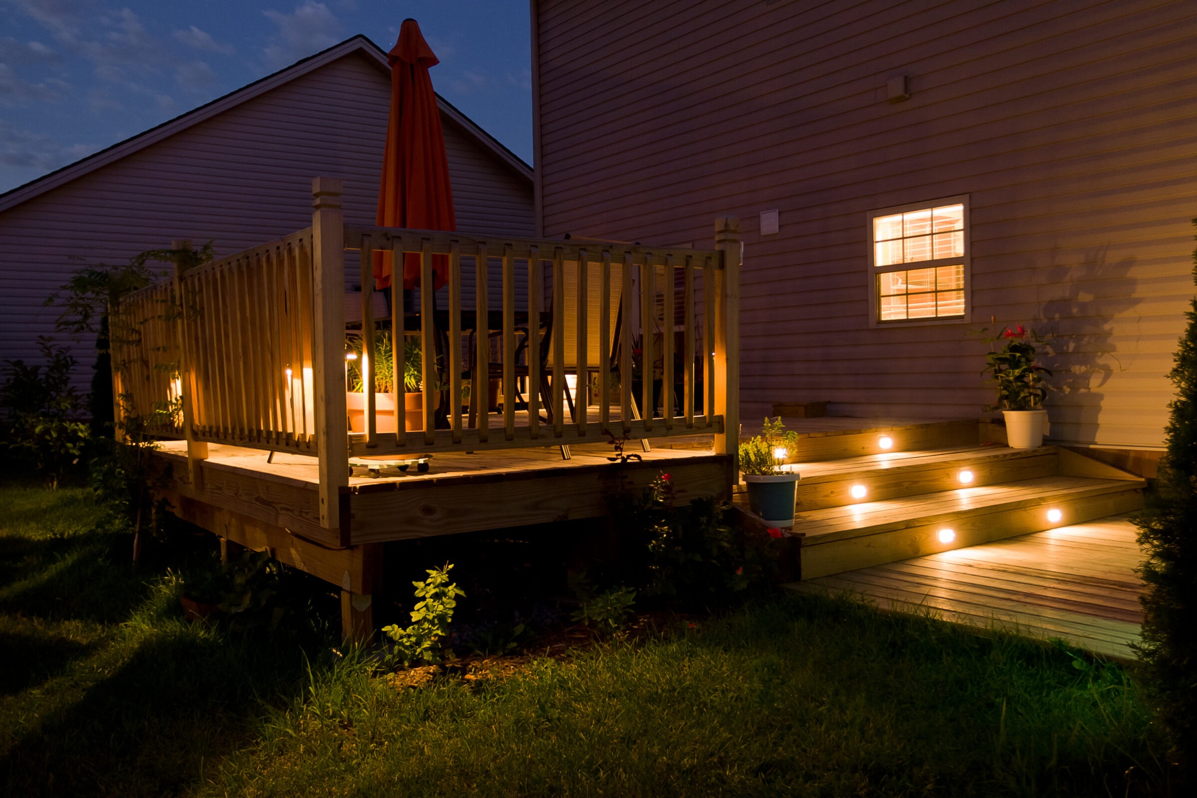 A cozy backyard deck at night, illuminated by soft lights, features potted plants and an umbrella under a clear evening sky.