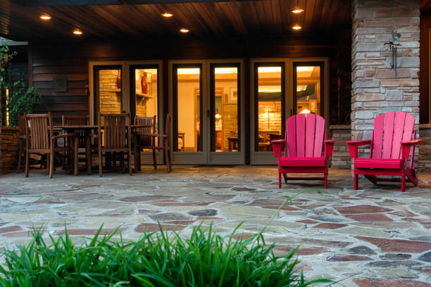 Two red Adirondack chairs on a stone patio, with wooden dining set nearby, viewed from a lush garden. Warm light emanates from interior.