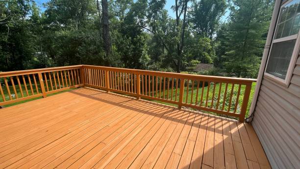 A spacious wooden deck overlooks a lush, green backyard with tall trees under a clear blue sky. Sunlight casts shadows on the deck.