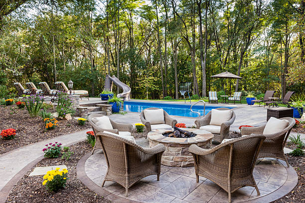 A serene backyard features a pool with a slide, surrounded by wicker chairs, colorful flowers, and tall trees, creating a tranquil retreat.
