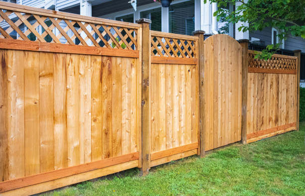 A wooden fence with lattice design tops and an arched gate stands near a house. Green grass surrounds the base of the fence.