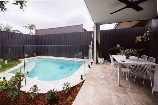 Modern backyard with a small pool, glass fence, and patio. Features a dining set, potted plants, and a dark wooden fence.