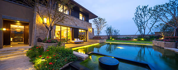 Modern house with illuminated windows, lush garden, and serene pool reflecting evening sky. Tranquil atmosphere enhanced by tasteful landscaping and ambient lighting.