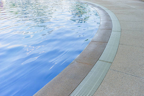 Curved swimming pool edge with clear blue water, surrounded by beige tiled flooring and metal drainage. No people or landmarks visible.