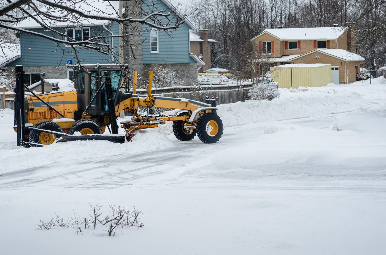 A yellow snowplow clears snow from a suburban street, surrounded by snow-covered houses and bare trees under a cloudy sky.